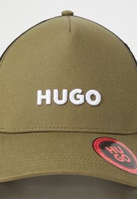 Olivgrüne Mütze mit strukturiertem Design, versehen mit weißer "HUGO"-Stickerei auf der Vorderseite und einem roten Markierungssticker an der Seite.