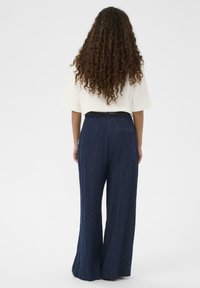 Oseba z dolgimi kodri, oblečena v belo kratkouhano srajco in temno modre široke hlače z visokim pasom, stoji s hrbtom obrnjena proti gledalcu.