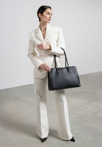 Black leather tote bag with textured finish, dual handles, and a structured shape, paired with a white tailored suit, and pointed black shoes.