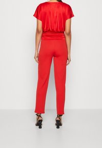 Rød satin top med korte ærmer og samlet talje, sat sammen med røde bukser i slim fit. Modellen bærer sorte hælede sandaler med runde detaljer.