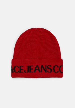 Bonnet rouge en tricot avec bord plié présentant un texte partiel noir "JEANS" sur le bord, sur un fond uni.