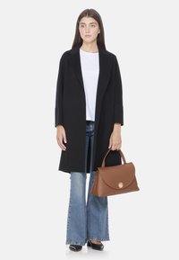 Manteau en laine noir avec un col à revers, associé à une chemise blanche et un jean évasé. Sac à main en cuir marron avec un détail de fermoir circulaire doré.