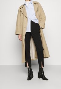 Impermeabile beige, camicia button-up azzurro chiaro, pantaloni neri su misura con spacchi laterali e stivaletti neri con tacco.