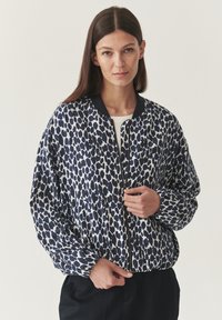 Vrouw met steil bruin haar, gekleed in een marineblauwe en witte stippen bomberjack met ritssluiting over een lichte top en zwarte broek.