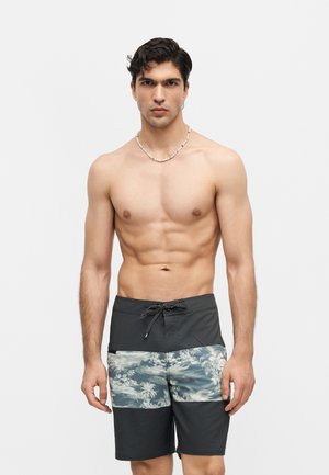 Άνδρας χωρίς μπλούζα με σκούρα μαλλιά που φοράει μαύρα και με σχέδια μαγιό, στέκεται μπροστά σε ένα απλό λευκό φόντο, κοιτώντας προς τα εμπρός.