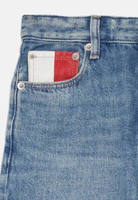 Lyse blå denimjeans har en rød og hvit lapp i baklommen, med sølvfarget tilbehør og synlige sømdetaljer.