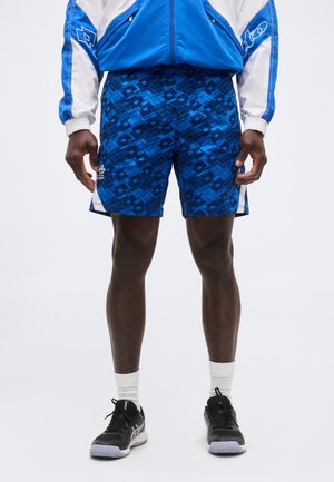 Mees, kes kannab siniseid geomeetrilise mustriga spordipükse, sinist ja valget jope, valgeid sokke ning musti spordijalatseid, seisab valge tausta ees.