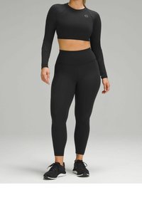 Černý sportovní crop top s dlouhými rukávy a legíny s vysokým pasem, obojí vyrobené z hladké, pružné tkaniny. Celkový vzhled doplňují pohodlné černé tenisky.
