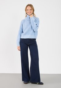 Ανοιχτό μπλε, υφασμάτινο πλεκτό πουλόβερ με ψηλό γιακά, συνδυασμένο με σκούρο μπλε, φαρδύ τζιν. Το μοντέλο στέκεται σε ουδέτερο φόντο.