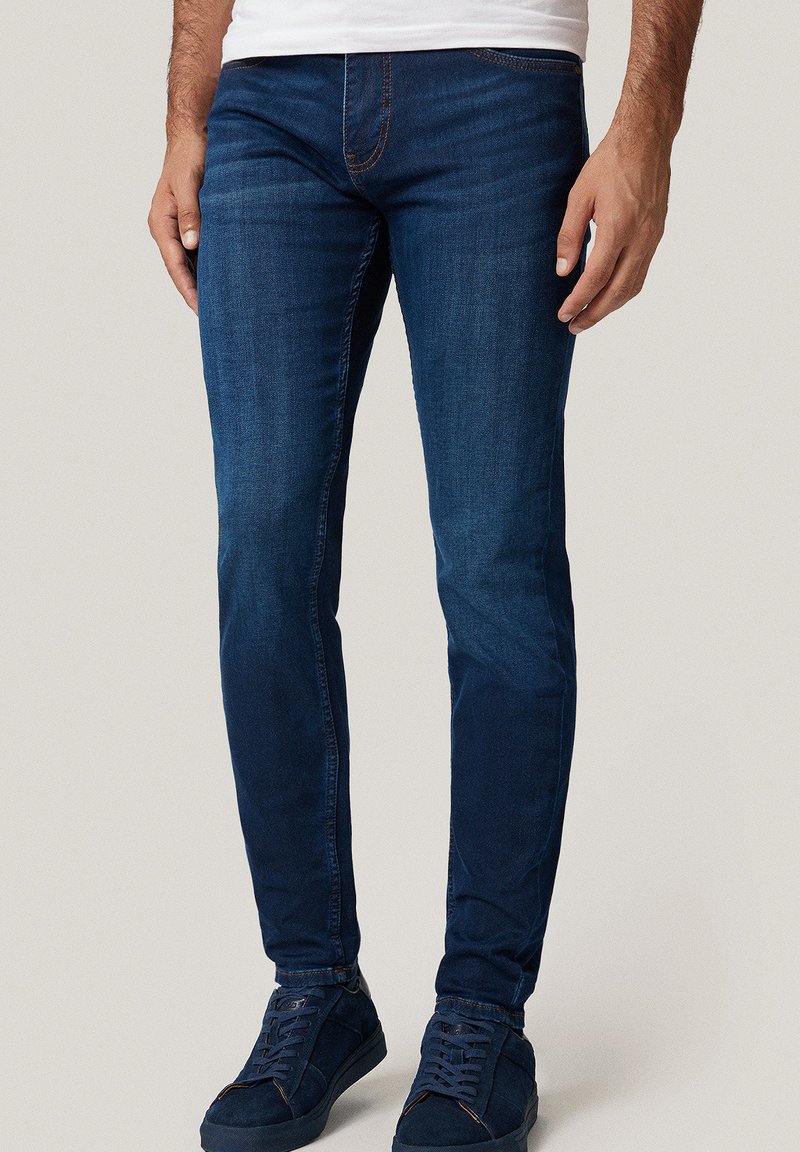 Mężczyzna w ciemnoniebieskich, slim-fit dżinsach i granatowych sneakersach, stojący na jednolitym jasnym tle.