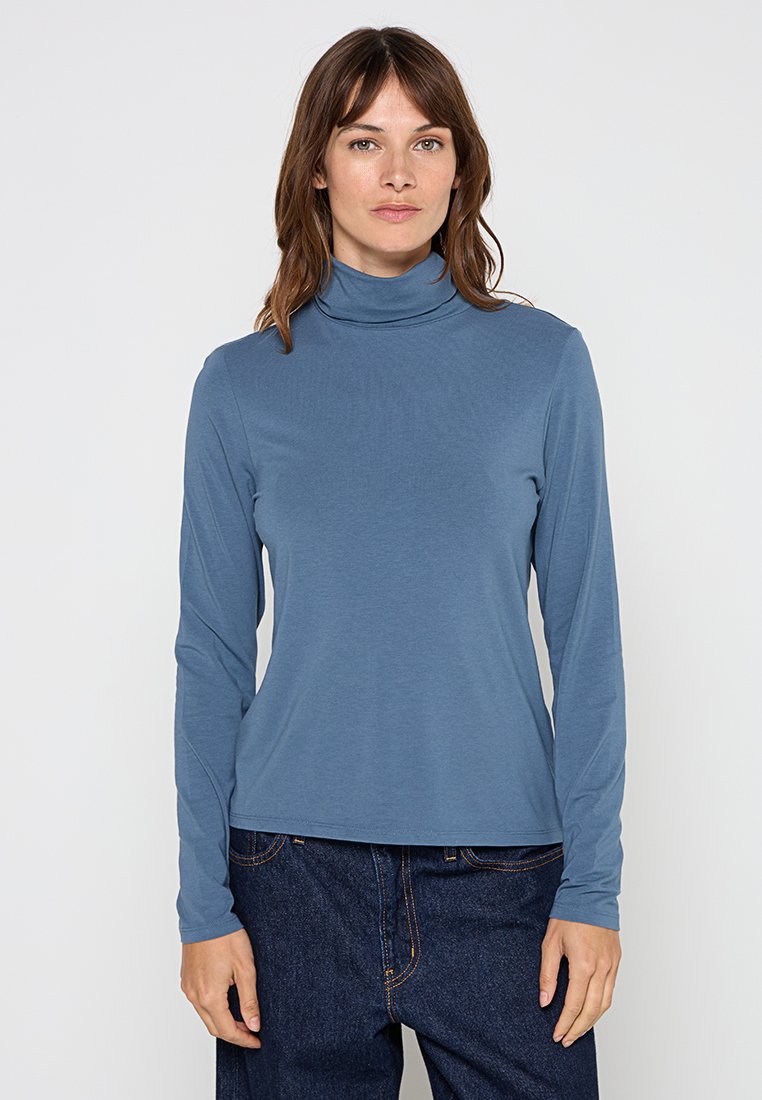 Langarm-Turtleneck-Shirt aus weichem blauen Stoff. Glatte Textur, enganliegendes Design und minimale Nähte. Kombiniert mit hoch geschnittenen dunkelblauen Jeans.