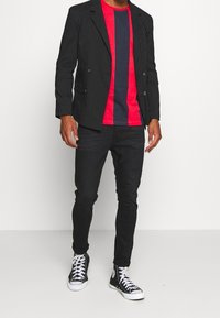 Un hombre se mantiene de pie con confianza en un blazer negro a rayas, camiseta a rayas roja y azul marino, jeans negros y zapatillas altas negras contra un fondo blanco.