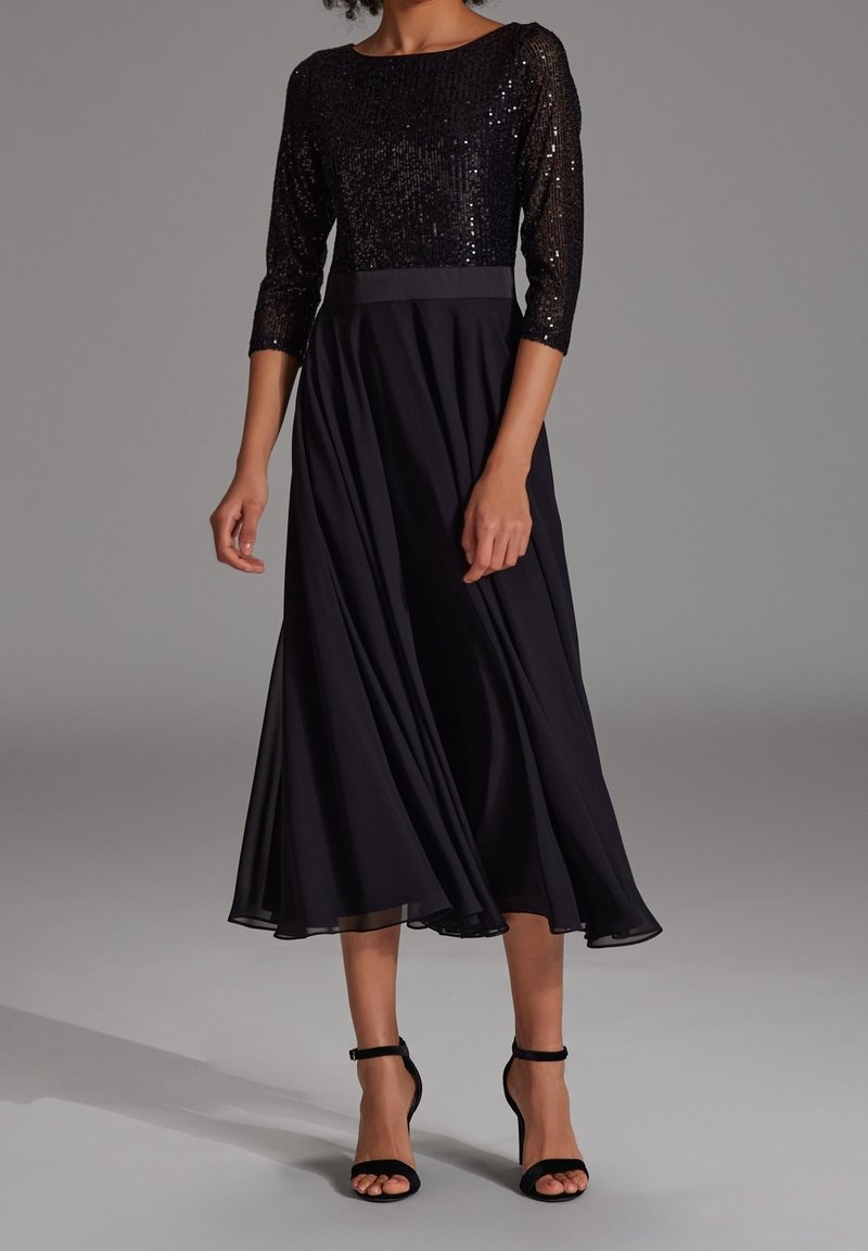 Femme portant une robe midi noire avec un corsage à sequins, des manches trois-quarts et une jupe fluide, assortie à des talons noirs à bride à la cheville.