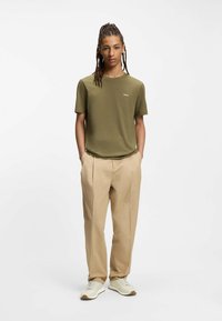 Camiseta de manga corta color verde oliva con un pequeño logo blanco, combinada con pantalones beige plisados y zapatillas blancas. Diseño sencillo, textura suave.