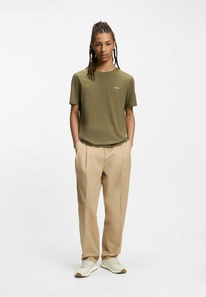 Olivgrünes Kurzarm-T-Shirt mit kleinem weißem Logo, kombiniert mit beigen plissierten Hosen und weißen Sneakers. Schlichtes Design, weiche Textur.