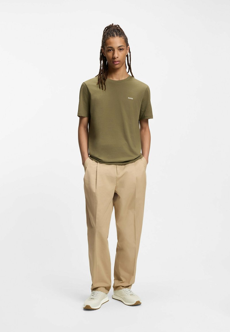 Camiseta de manga corta color verde oliva con un pequeño logo blanco, combinada con pantalones beige plisados y zapatillas blancas. Diseño sencillo, textura suave.