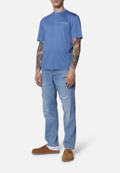 Maglietta blu a maniche corte con scollatura rotonda e un piccolo logo bianco sul petto, abbinata a jeans chiari strappati e zoccoli marroni.