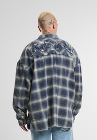 Chemise en flanelle à carreaux bleu marine et beige, avec une coupe décontractée, des détails usés et un logo brodé "KANU" au dos. Manches à boutons.