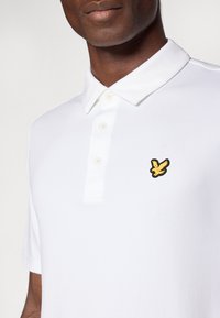 Balta polo marškinėliai su apykakle, penkių sagų užsegimu ir geltonai juodu siuvinėtu logotipu ant krūtinės. Sklandžiai tekstūruotas audinys.