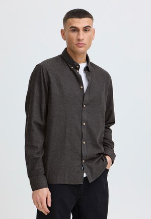 Jeune homme aux cheveux courts portant une chemise marron foncé boutonnée par-dessus un t-shirt blanc et un pantalon noir, posant avec une main dans la poche.