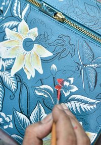 Blauwe leren oppervlak met bloemontwerpen. Een hand houdt een penseel vast en voegt rode accenten toe aan een bloem naast bladeren en een vlinderillustratie.