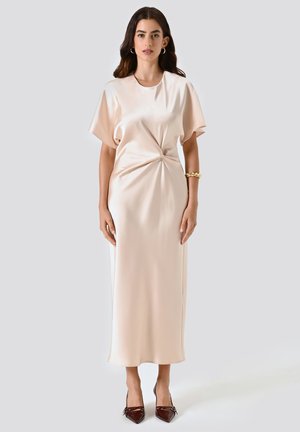 Una donna con lunghi capelli scuri indossa un midi dress beige in satin con maniche corte, un dettaglio a nodo laterale, un bracciale d'oro, orecchini a cerchio e tacchi a punta scuri.
