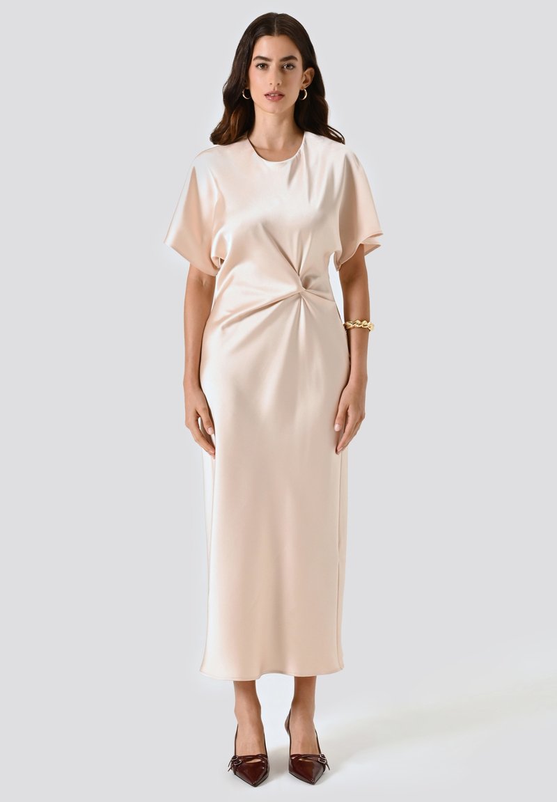 Una donna con lunghi capelli scuri indossa un midi dress beige in satin con maniche corte, un dettaglio a nodo laterale, un bracciale d'oro, orecchini a cerchio e tacchi a punta scuri.