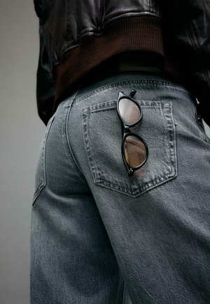 Oseba, oblečena v izprane modre kavbojke in črno usnjeno jakno, z za kavbojkami zataknjenimi črnimi sončnimi očali.