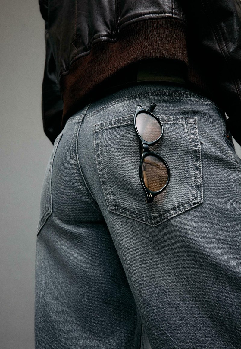 Personne portant un jean bleu délavé et une veste en cuir noire avec des lunettes de soleil noires accrochées à la poche arrière du jean.