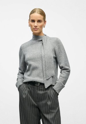 Grauer Strickpullover mit hohem Kragen und angehängtem Bindebanddetail, kombiniert mit schwarzen Nadelstreifenhosen. Glatte Textur und lässiger Schnitt.