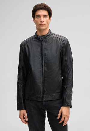 Mann steht vor einem schlichten Hintergrund, trägt eine schwarze Lederjacke mit Reißverschluss und gesteppten Schultern sowie eine schwarze Jeans, schaut nach vorne.