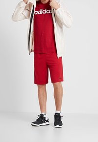 Muž v červeném tričku a kraťasech značky Adidas, černo-bílých teniskách, upravuje bílou bundu s černými zipy.