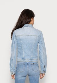 Žena s dlouhými tmavými vlasy, oblečená v světle modré džínové bundě a odpovídajících džínech, stojící zády k objektivu na jednoduchém pozadí.