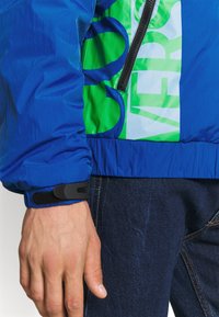 Blå lättviktig jacka med gröna och blå paneler; har en dragkedjeficka och elastiska ärmslut med ett justerbart svart band.