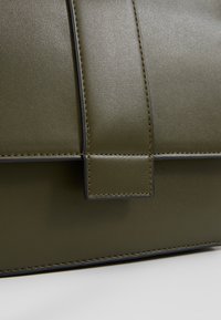 Acercamiento a un bolso de cuero color verde oliva con costuras visibles y detalles de solapa sobre un fondo blanco.