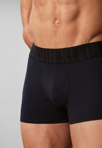 Ανδρικός κορμός με μαύρα μπόξερ Calvin Klein με ορατό λογότυπο στη μέση, σε απλό γκρι φόντο.