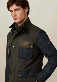 Mladic nosi zelenu i crnu jaknu s više džepova na zakopčavanje i oznakom "Hackett Velospeed" na prsima.