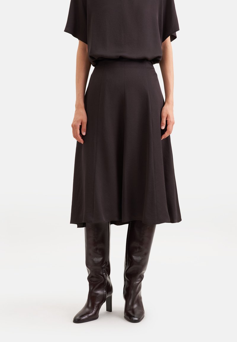 Femme portant une jupe midi noire associée à des bottes noires à talons hauts montant jusqu'aux genoux sur un fond blanc uni.