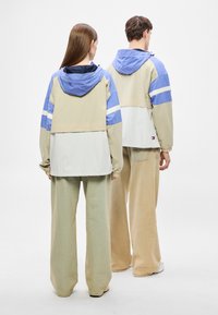 Könnyű, színblokkokkal díszített anorák bézs, kék és fehér panelekkel. Kapucnival, elasztikus mandzsettákkal és bő, zöld kordbársony nadrággal rendelkezik.
