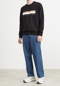 Svart sweatshirt med horisontell färgblock i beige, vitt och grått. Matchas med ljusblå jeans och vita sneakers, står på ett neutralt golv.