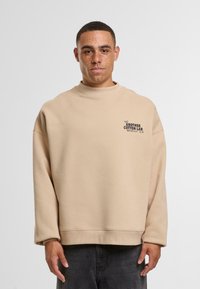 Felpa oversize beige con colletto a girocollo alto e maniche lunghe. Presenta una stampa logo frontale e polsini a costine.