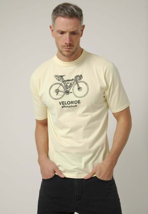 Hombre con el pelo corto vistiendo una camiseta color crema con un dibujo detallado en negro de una bicicleta y el texto “VELORIDE altonadock.”