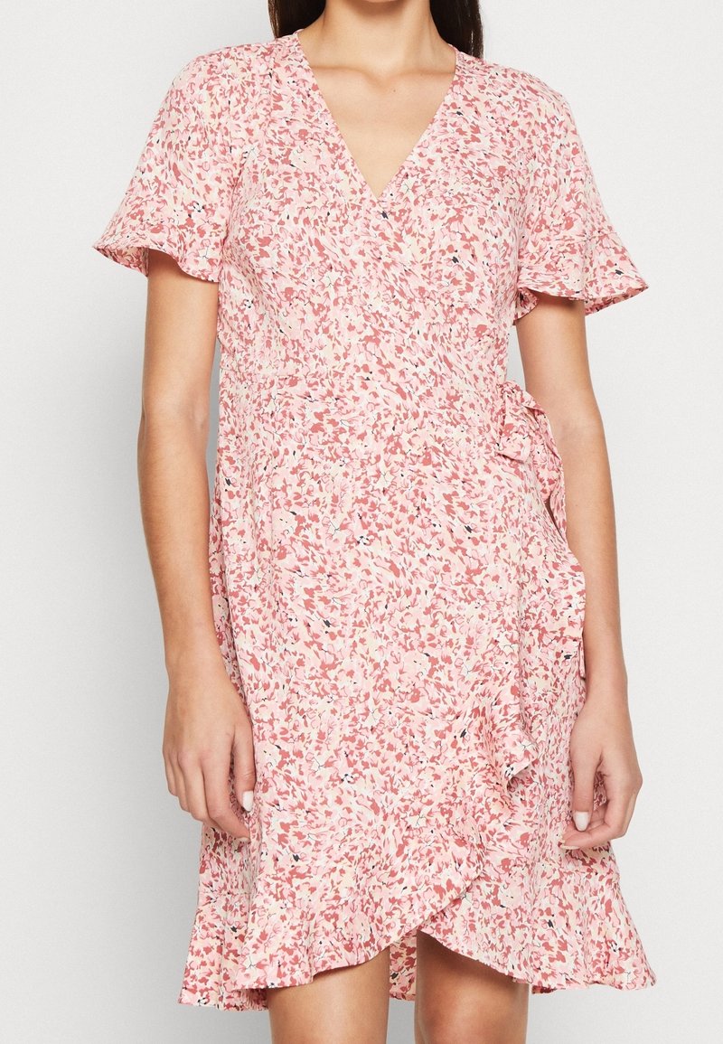Femme portant une robe portefeuille rose à manches courtes avec un petit imprimé floral et un ourlet et des manches volantés.