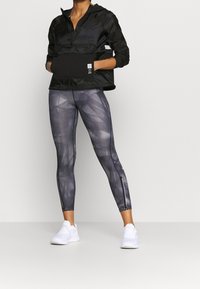 Czarna kurtka z kapturem i półzamkiem, z przednią kieszenią; szare legginsy z abstrakcyjnym wzorem i suwakami przy kostkach; białe buty sportowe.