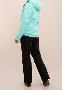 Une veste vert menthe avec capuche et un pantalon noir. La veste possède une texture lisse, tandis que le pantalon est en tissu robuste et mat.