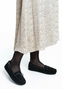 Zwarte suede moccasin pantoffels met veterdetails, gedragen met zwarte panty en een gemêleerde beige rok met kleine zwarte stippen.