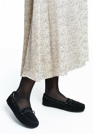 Mocasines de ante negro con detalle de cordones, usados con medias negras y una falda beige con estampado que presenta pequeños puntos negros.