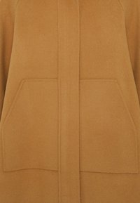 Cappotto di lana beige con una trama morbida, dotato di chiusura con patta frontale e due grandi tasche applicate. Design semplice, senza decorazioni.