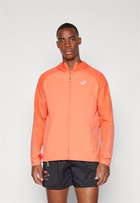 Veste à capuche zippée en orange vif, en matériau léger, avec un logo sur la poitrine et un design bicolore avec des poches latérales.