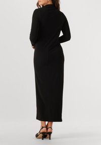 Une femme portant une longue robe noire ajustée avec des manches longues et des sandales à talons noires, se tenant les mains sur les hanches en tournant le dos.